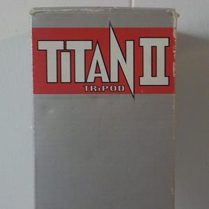 May include: A gray box with the words "TITAN II TRIPOD" in white letters on a red background. The box is rectangular and appears to be made of cardboard. The text is centered and easy to read.
