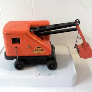 May include: A vintage orange and black metal toy crane with a mechanical shovel arm. The main body features a small window and a circular logo with the text Structo Construction. The toy is mounted on four black wheels and has a black boom arm.