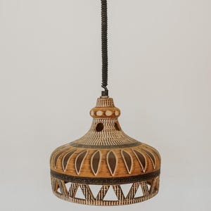 May include: A ceramic pendant light with a tan and dark brown glaze. The bell-shaped shade features carved petal patterns, circular cutouts near the top, and a geometric openwork border at the base. It hangs from a black cord and ceiling canopy.