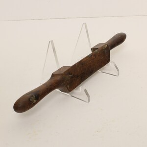 May include: A vintage wooden hand plane with two wooden handles. The plane is resting on a clear acrylic stand.