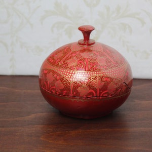 Op de afbeelding: Een ronde, rode decoratieve doos met een deksel. De doos is versierd met ingewikkelde gouden bloempatronen. Het deksel heeft een kleine, afgeronde knop. De doos staat op een donkerbruin houten oppervlak.