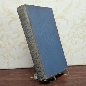 May include: An old book with a blue cover and an embossed floral pattern. The spine is grey with gold lettering. The book is displayed on a clear acrylic stand, highlighting its vintage look and historical appeal.