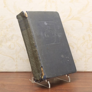 Puede incluir: Un libro antiguo con una cubierta azul oscuro, exhibido en un soporte de acrílico transparente. El lomo del libro muestra desgaste, con daños visibles en los bordes. La cubierta presenta un diseño en relieve descolorido.