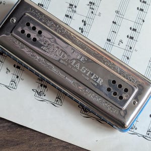 Puede incluir: Una armónica plateada vintage con la inscripción "THE BANDMASTER" y "MADE IN GDR" se muestra. La armónica tiene un borde azul y está colocada sobre una hoja de partituras. El instrumento presenta múltiples agujeros y grabados decorativos.