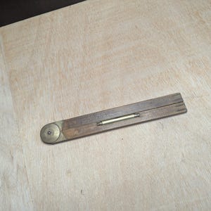 Puede incluir: Un calibrador de bisel de madera vintage con accesorios de latón. Esta herramienta se utiliza para medir y transferir ángulos. La madera tiene un aspecto oscuro y envejecido, lo que sugiere que es una herramienta antigua de carpintería.