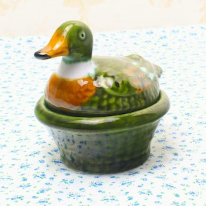 以下が含まれることがあります： 緑色の頭、オレンジ色のくちばし、白い首を持つ陶器製のアヒルの置物。アヒルは緑色のボウル型の台座の上に座っています。アヒルの体は緑、オレンジ、茶色の混合色です。この置物は装飾品である可能性が高いです。