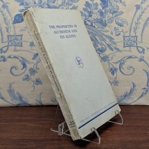 May include: An old book, "The Properties of Aluminium and Its Alloys", is displayed on a clear acrylic stand. The cover is pale with blue text and a circular logo. The background shows a blue and white floral design, typical of vintage decor.