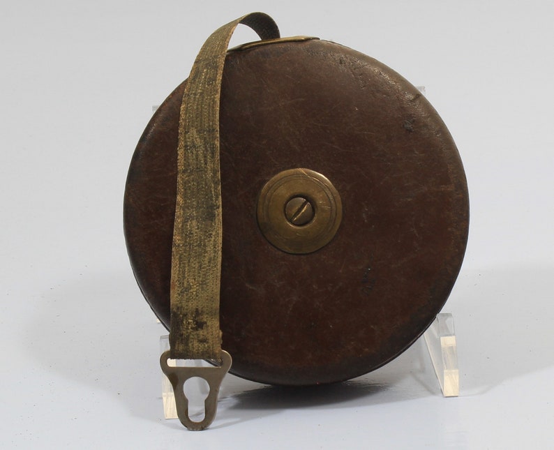 May include: A vintage brown leather tape measure with a brass winding mechanism and a metal hook. The tape measure is rolled up and secured with a brown leather strap.