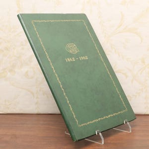 May include: A green book with gold trim and the text "1862-1962" is displayed on a clear acrylic stand. The book has a gold border and a logo in the center. The background is a floral pattern.