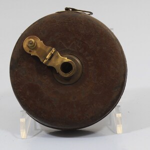 May include: A vintage wooden tape measure with a brass winding mechanism and a leather case. The case is brown and has a worn look. The brass mechanism has a small handle and a hole in the center.