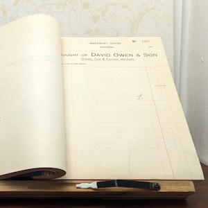 Op de afbeelding: Een openstaand notitieboek met gelinieerde pagina's, met de tekst "FOUGHT OF DAVID OWEN & SON" en "Grocers, Corn & Provision Merchants". Het notitieboek wordt opengehouden door een houten boekenstandaard met een zwart-witte metalen clip.