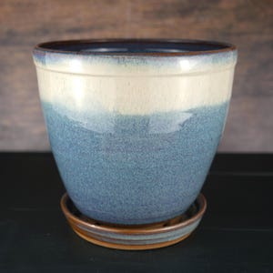 May include: A ceramic plant pot with a rounded shape, featuring a two-tone glaze with a creamy white upper section and a speckled blue lower section. The pot includes a matching circular saucer base with a brown rim and interior.