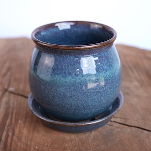 May include: A round ceramic planter with a glossy finish in shades of deep blue and teal, featuring a dark brown rim. The planter sits on a matching circular saucer, creating a cohesive set for holding small plants or decorative items.