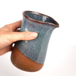 May include: A ceramic pitcher featuring a speckled blue and grey glaze on the upper portion and a solid, unglazed brown clay base. The vessel has a flared rim with a distinct pouring spout and a smooth, tapered body design for liquid pouring.