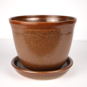 May include: A brown ceramic plant pot with a speckled finish and a matching circular saucer. The pot features a wide, flared rim and a smooth, glazed surface that transitions from a darker tone at the base to a lighter brown at the top edge.
