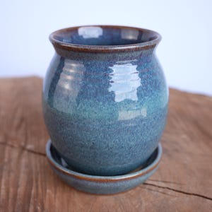 May include: A round ceramic planter with a glossy blue and teal glaze, featuring a smooth, bulbous body and a flared rim. The planter includes a matching circular saucer base, both finished with a dark brown edge along the top and bottom rims.