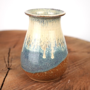 May include: A ceramic vase with a flared rim, featuring a three-tone glaze. The top section is a creamy off-white, transitioning into a mottled blue drip pattern that overlays a textured, earthy brown base at the bottom of the vessel.