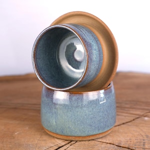May include: Two ceramic cups with a blue and grey mottled glaze and exposed brown clay rims. One cup is positioned upright, while the second cup is inverted and balanced on top of the first, revealing the smooth interior and the circular base.