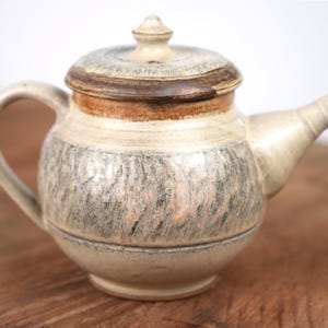 May include: A ceramic teapot with a rounded body and a matching lid. The surface features a cream-colored base with textured, vertical brushstrokes in shades of gray and brown. It includes a curved handle and a tapered spout extending to the right.