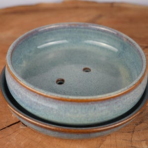 May include: A round ceramic soap dish with a light blue and grey speckled glaze. The dish features a raised rim with a brown edge and two drainage holes in the center. It sits nested inside a matching circular saucer with a dark brown rim.