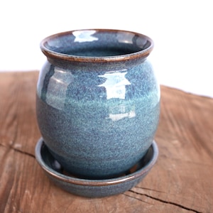 May include: A round ceramic planter with a glossy blue and teal speckled glaze. The pot features a dark brown rim and sits on a matching circular saucer. The surface of the pottery shows variations in color tone, creating a textured appearance.