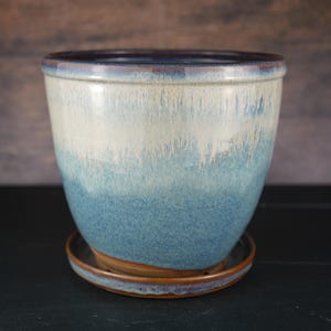May include: A ceramic plant pot with a matching saucer, featuring a glossy glaze that transitions from a deep blue at the base to a lighter, creamy white at the rim, with a dark brown rim and base edge visible on the saucer.