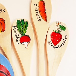Può includere: Quattro cucchiai di legno dipinti con diverse verdure e frutta, tra cui peperoni, carote, ravanelli e fragole. I cucchiai sono disposti a ventaglio su una superficie bianca. Uno sfondo con una pietra dipinta con una coccinella.