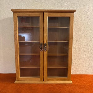 May include: A wooden cabinet with two glass doors and multiple shelves. The cabinet is made of light-colored wood and features ornate metal handles. The interior is visible through the glass doors, showcasing the shelves. The cabinet is designed for storage and display.