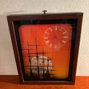 May include: A framed wall clock with a nautical theme. The clock face is white with black Roman numerals. The background features a silhouette of a ship against a sunset. The frame is dark brown.