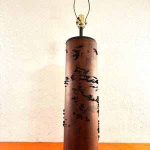 May include: A cylindrical table lamp with a dark brown wooden shade featuring cut-out designs. The lamp has a brass-colored bulb holder and a dark base. The lamp's height is approximately 24 inches.