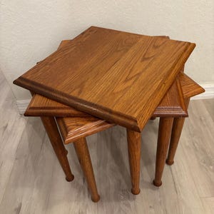 May include: A set of three nested tables made of medium-toned wood with a visible grain pattern. Each table features a square top with beveled edges and four tapered cylindrical legs. The tables are stacked together in a tiered arrangement.