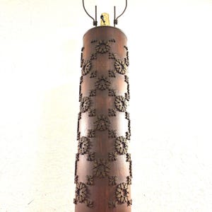 May include: A vintage table lamp with a cylindrical wooden base. The base is adorned with repeating floral metal accents. The lamp has a metal frame and a light bulb. The lamp is plugged into an electrical outlet.