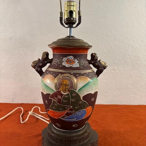 May include: A brown ceramic table lamp with gold accents and a painted scene of a person in a robe. The lamp has two handles and a round base.
