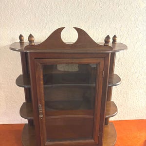 May include: A dark wood curio cabinet with a glass door and multiple shelves. The cabinet has a decorative top with a crescent-shaped cutout and small finials. The shelves are curved, and the cabinet sits on a rounded base.