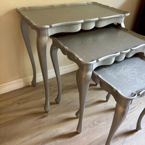May include: A set of three silver nesting tables with scalloped edges and ornate legs. The tables are arranged in a stack, with the largest table on the bottom and the smallest table on top.
