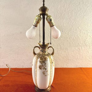 May include: Antique white ceramic lamp with a floral design and brass accents. The lamp features two light bulb sockets and a decorative base. The lamp has a vintage aesthetic and is suitable for home decor.