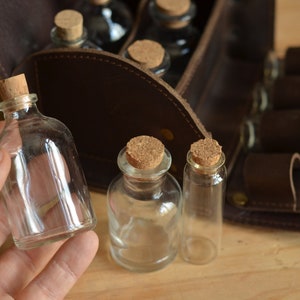 Peut inclure: Un ensemble de petites bouteilles en verre avec des bouchons en li&egrave;ge, dispos&eacute;es dans un &eacute;tui de transport en cuir marron. Les bouteilles sont transparentes et vides.