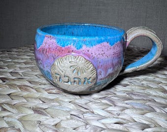 Op de afbeelding: Een keramische mok met een blauwe binnenkant en een handvat. De buitenkant heeft een mix van blauwe, roze en paarse tinten. De mok heeft een cirkelvormig embleem met Hebreeuwse tekst. De mok staat op een geweven oppervlak.