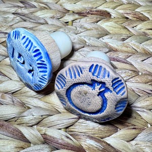 May include: Two ceramic bottle stoppers with blue and white designs. One stopper has a blue design with a circular pattern, and the other has a blue design with a curved shape. The stoppers have wooden and white plastic components.