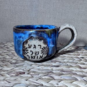 May include: A handmade ceramic mug with a blue glaze and a white handle. The mug features a decorative emblem with text in a foreign language. The mug is sitting on a woven surface.