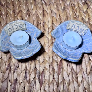 May include: Two light blue ceramic candle holders with irregular, organic shapes. Each holder features a small, light blue tea light candle and a rectangular plaque with Hebrew text. The left plaque reads Shalom, and the right plaque reads Shabbat.