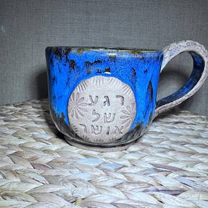 May include: A ceramic mug with a blue glaze and a textured handle. The mug features a circular emblem with Hebrew text and floral designs. The mug is set on a woven surface.