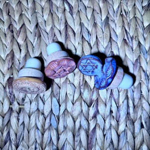 May include: Three bottle stoppers with light-colored wooden bases and white silicone tips. Two stoppers feature round, tan-colored ceramic tops with embossed star designs, while the third has a blue, textured, irregular ceramic top.