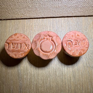 May include: Three circular ceramic objects with a glossy, light brown finish. The left object features Hebrew text, the center object displays an embossed pomegranate design, and the right object features Hebrew text. Each piece has a textured edge.