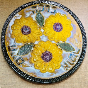 May include: A ceramic plate with a black and white border and a design of three yellow flowers with brown centers. The plate has the Hebrew words "L'Chaim" written in black on the top and "Mazal Tov" written in black on the bottom.