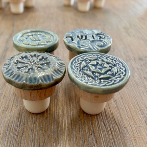 May include: Four decorative bottle stoppers with ceramic tops in shades of green and brown. Each stopper features a unique embossed design, including floral and Celtic knot patterns. The stoppers are made of wood and ceramic.