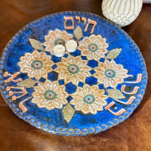 May include: A blue ceramic plate with a white floral design and the Hebrew word "Shalom" in the center. The plate has a raised rim and a scalloped edge.