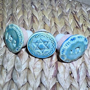 May include: Three bottle stoppers with light blue ceramic tops and wooden bases. The left stopper features Hebrew lettering, the center stopper displays a six-pointed star with a braided border, and the right stopper features a single Hebrew character.