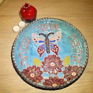 May include: A blue ceramic plate with a butterfly design and Hebrew writing. The plate has a floral border and is decorated with red, white, and blue accents.