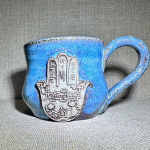 May include: A blue ceramic mug with a white hamsa hand design. The hamsa hand has Hebrew writing on it.